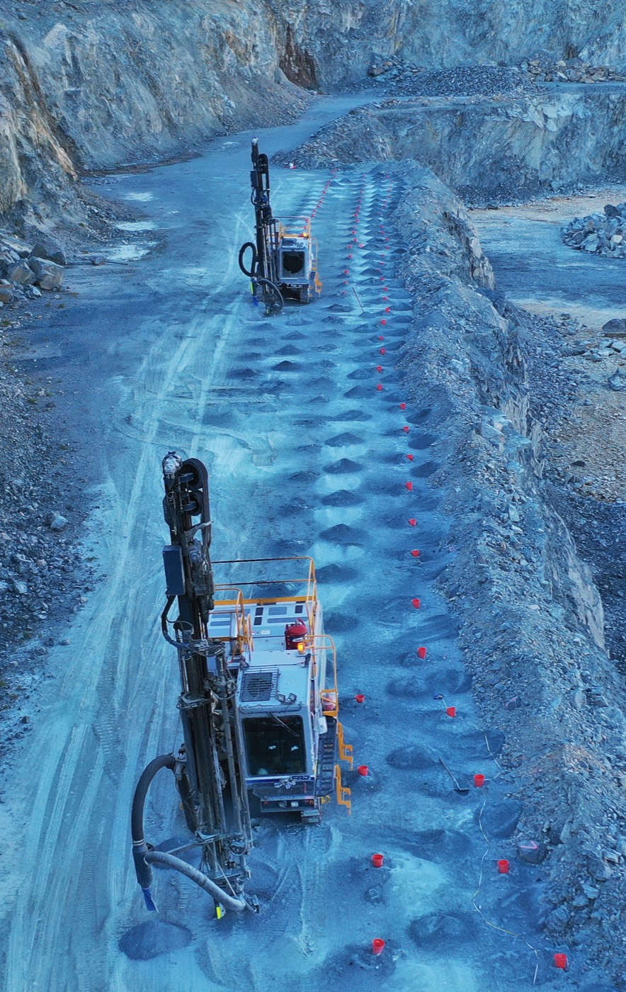 Aerial view of two drill rigs working along a drilled line on a quarry bench