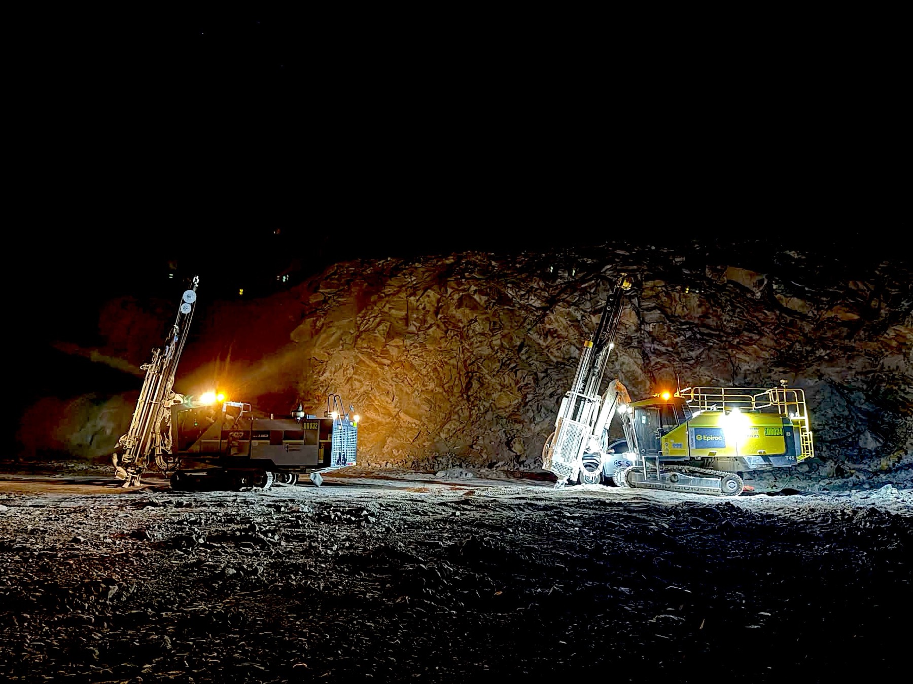 Two drill rigs working under lights against a rock face at night