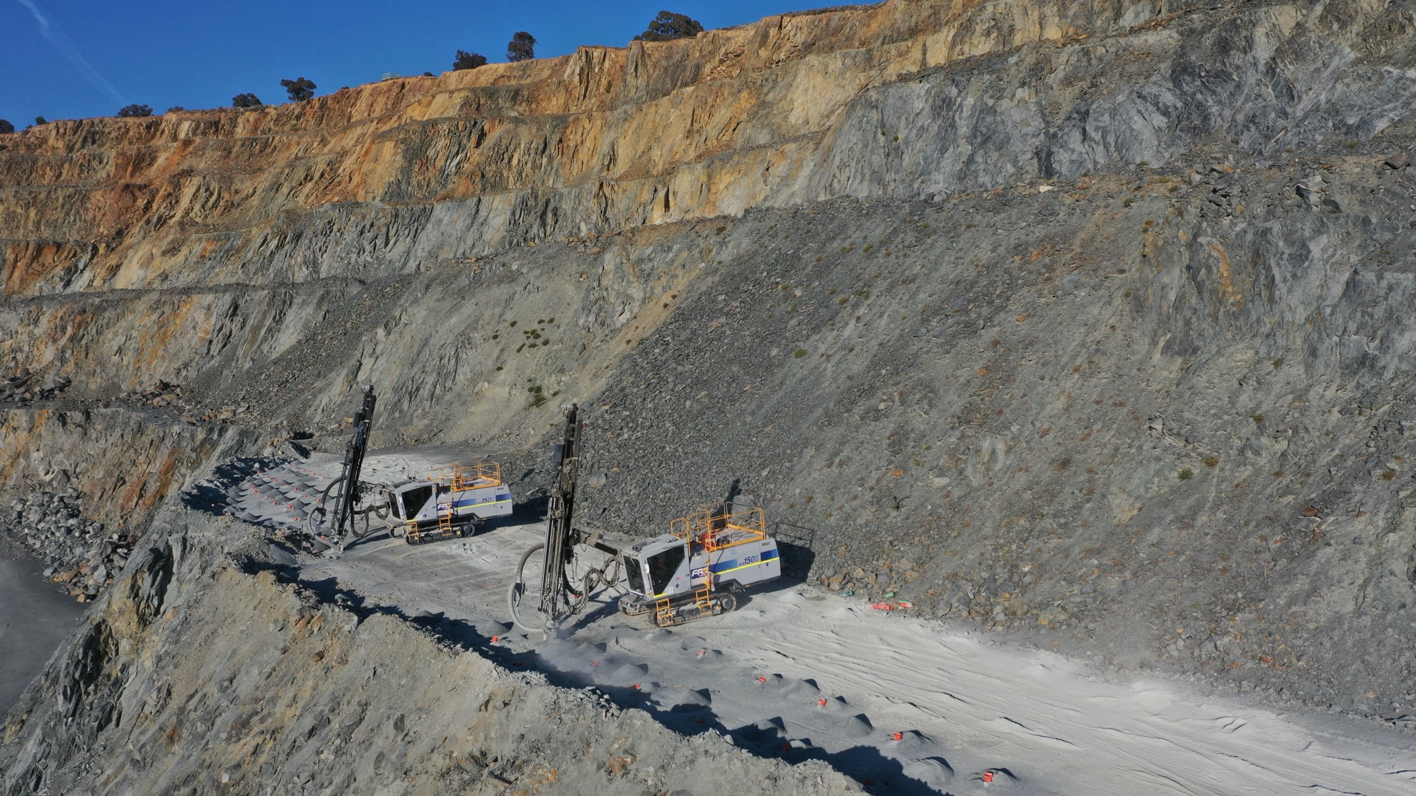Two drill rigs working a drilled bench beneath a high quarry wall